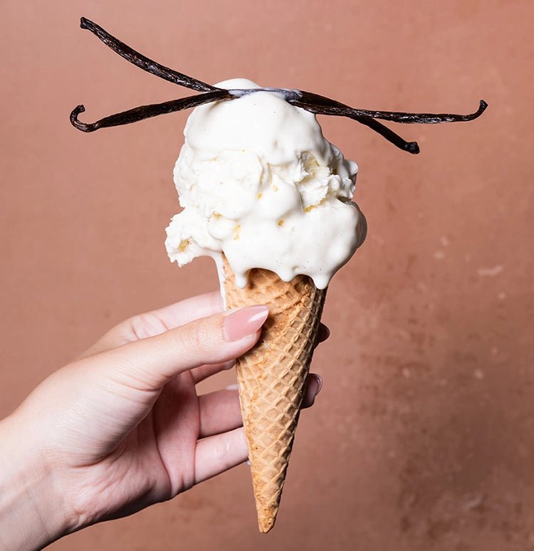 A hand holding a vanilla ice cream cone with two vanilla bean slices on top, set against a sol-id terracotta background.