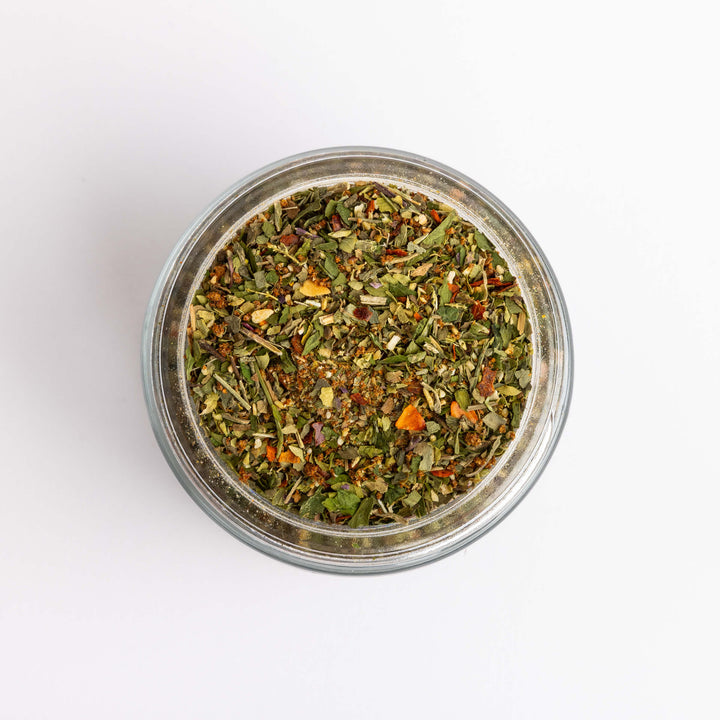 Top-down view of a clear glass jar filled with a loose spice blend containing green, brown, and orange dried botanicals, set against a plain white background.